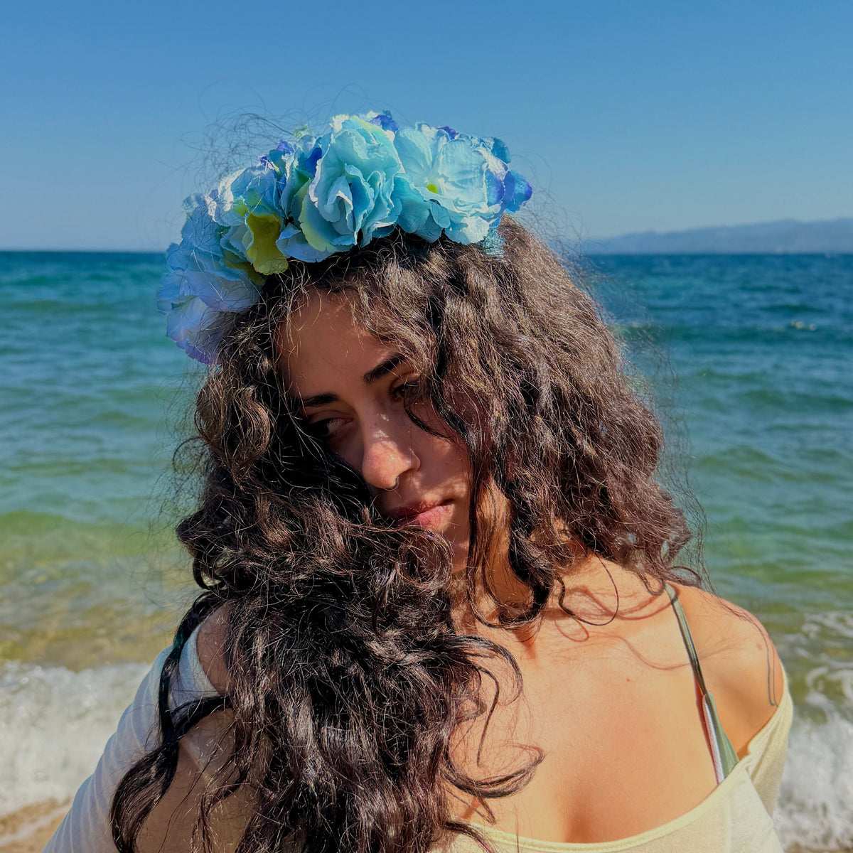 A woman with long, curly dark hair wearing a light blue flower crown stands on a beach with the ocean and blue sky in the background.