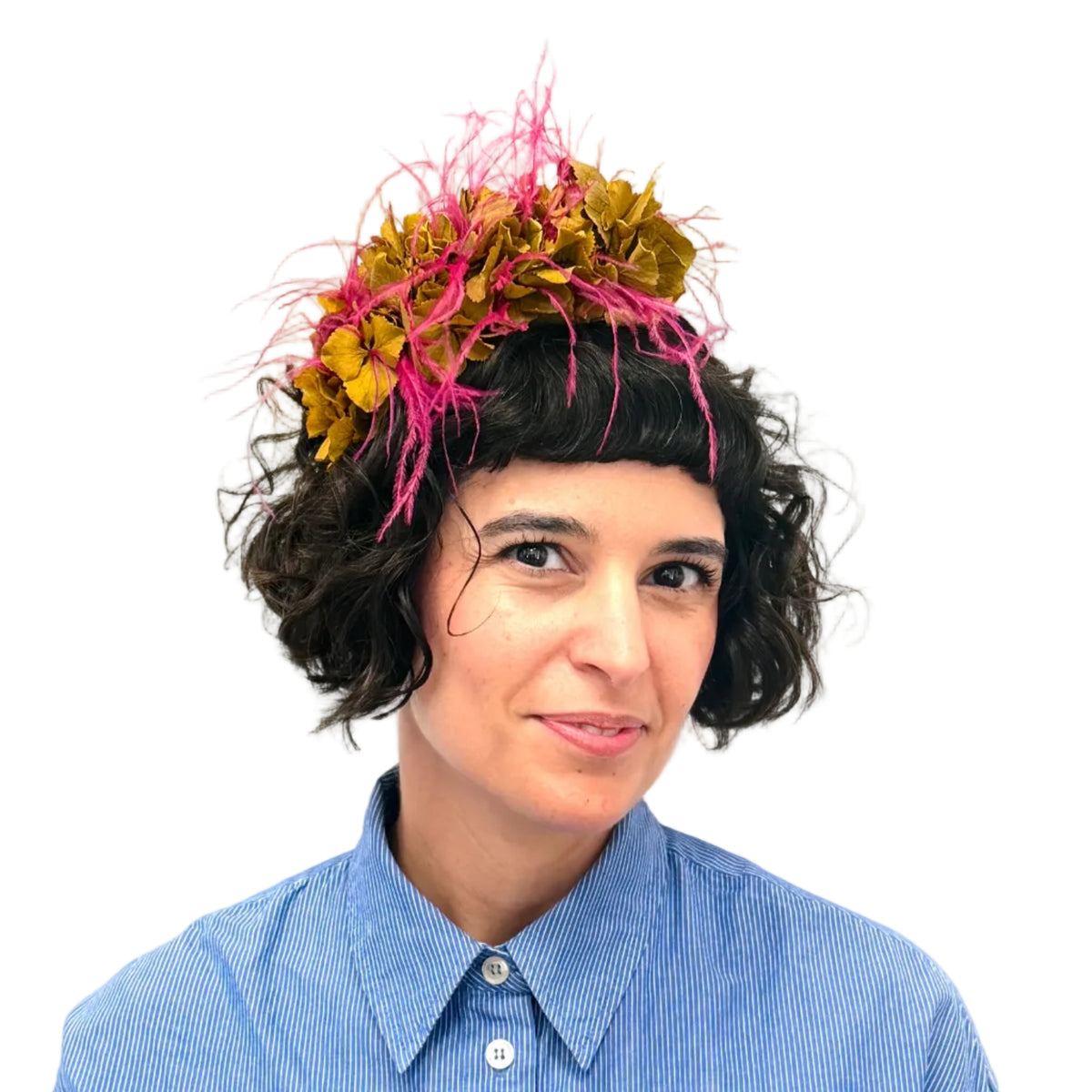 A woman with short dark curly hair wearing a vibrant pink and yellow dried flower headpiece, paired with a light blue button-up shirt, set against a plain white wall with a colorful botanical painting partially visible above.