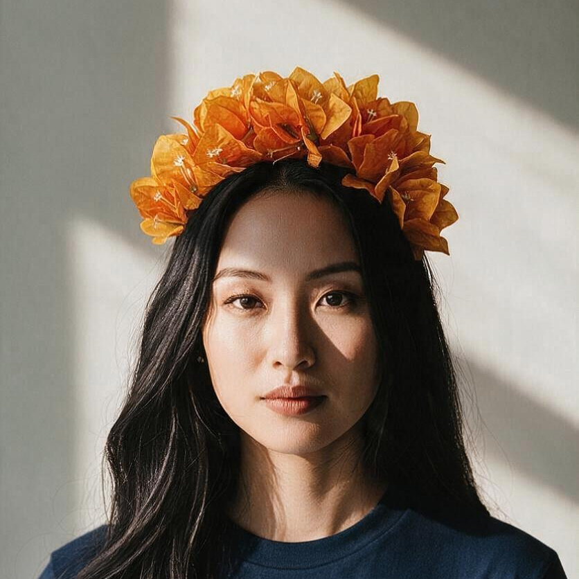 A woman with long dark hair wearing a voluminous orange flower crown, photographed against a warm-toned wall with dramatic diagonal sunlight and shadows.