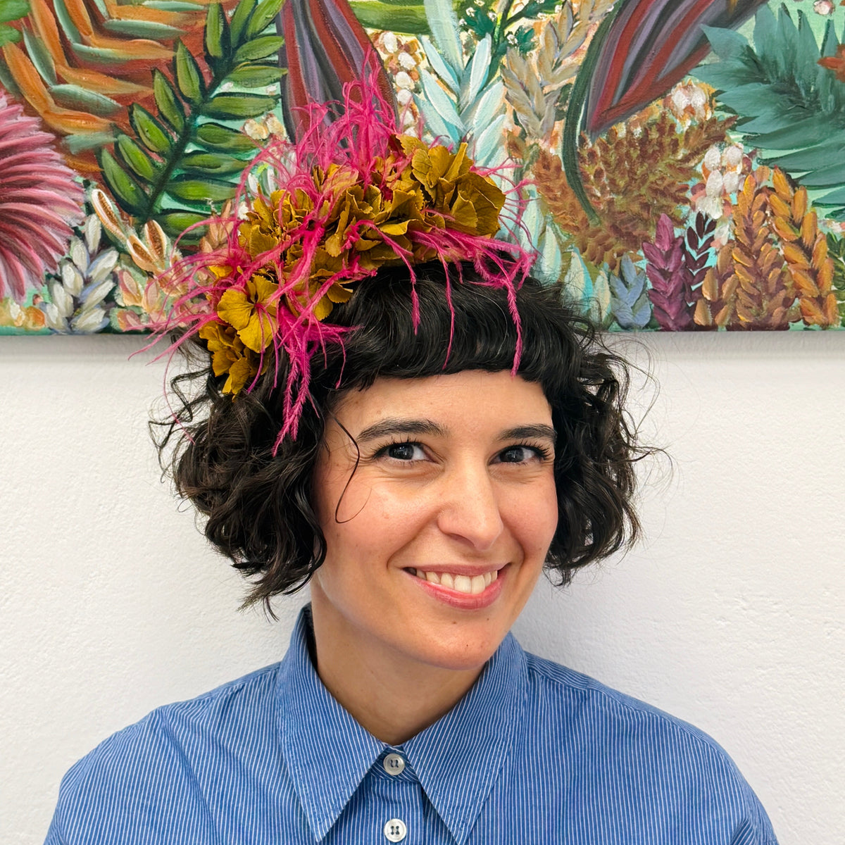 A smiling woman with short curly dark hair wears a colorful floral headpiece with bright pink, yellow, and orange flowers. She is standing in front of a white wall with a large tropical artwork hanging above her head.