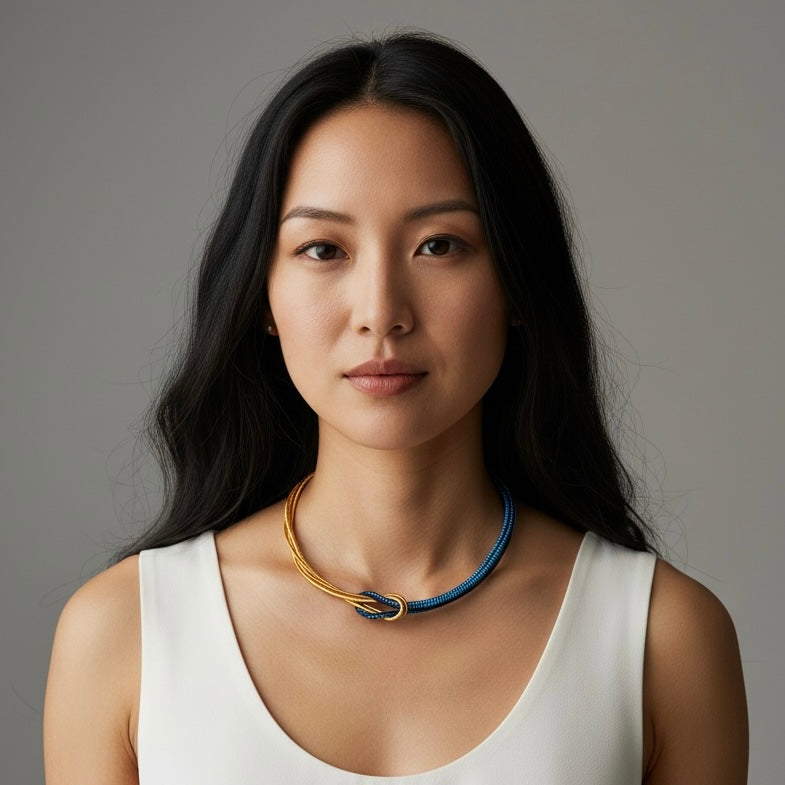 A woman with long black hair wearing a white sleeveless top and a choker necklace with gold and blue braided details, set against a solid gray background.