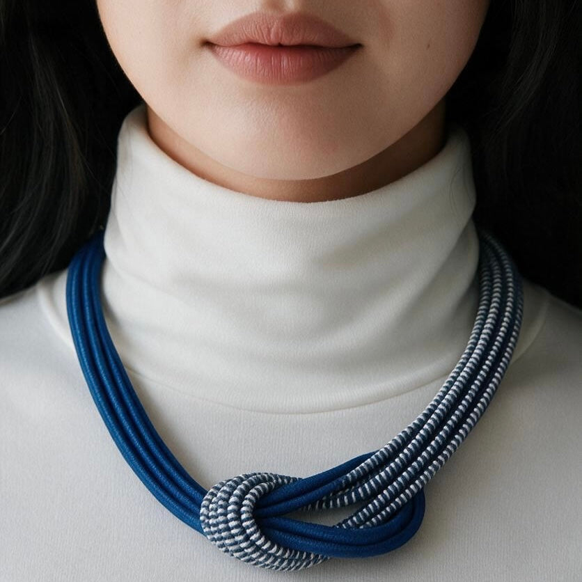 A close-up of a model wearing a multi-strand statement necklace with a knot detail. The necklace features dark blue cords mixed with silver and white striped cords, styled over a white turtleneck sweater.