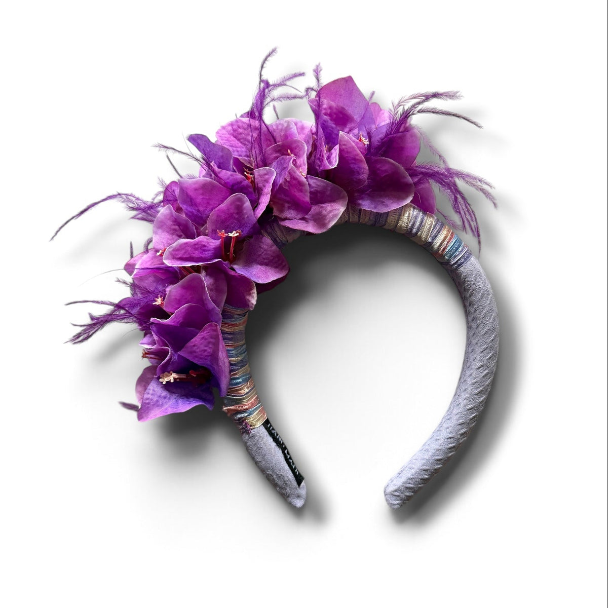 A violet fabric headband decorated with a cluster of purple artificial flowers and wispy purple feathers, shown on a plain white background.